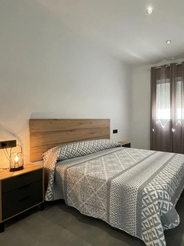 Dormitorio con cama doble y ropa de cama gris, lámpara de noche y cortinas en la ventana