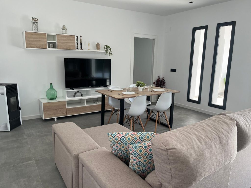 Sala de estar y comedor con sofá gris, mesa de comedor con sillas blancas y televisor en pared blanca